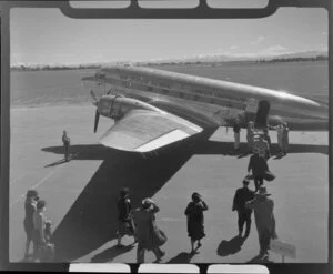 Harewood, Christchurch, New Zealand National Airways Corporation DC3 on runway, with passengers embarking through rear doorway of plane via stairs, snow covered mountains beyond