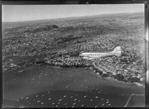 New Zealand National Airways Corporation (NAC) Flagship Dakota aircraft in flight over Auckland, showing boats in the harbour