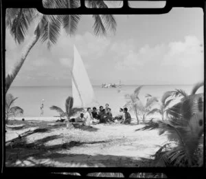 Local boys relax on beach, Akaiami, Aitutaki, Cook Islands, with TEAL (Tasman Empire Airways Limited) Flying boat in the background