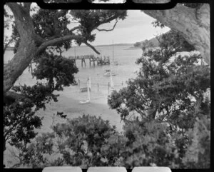 Beach scene at Paihia, Bay of Islands, showing bathers, boats and wharf through the trees