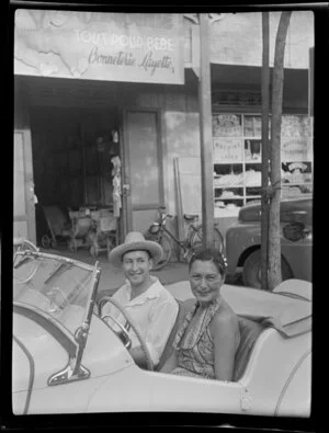 Mr Neil Hilton and Suzanne, sitting in MG convertable motor car, outside general store, Papeete, Tahiti