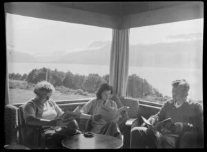 Interior view of unidentified group reading in the lounge of a lodge at Lake Ohau, Waitaki District, Canterbury Region, showing mountains through the windows