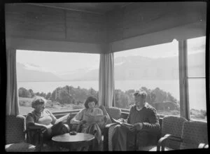 Interior view of unidentified group reading in the lounge of a lodge at Lake Ohau, Waitaki District, Canterbury Region, showing mountains through the windows