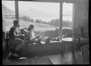 Interior view of a couple in the lounge of a lodge at Lake Ohau, Waitaki District, Canterbury Region, showing mountains through the windows