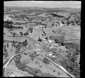 Waimauku, Auckland, showing intersection of State Highway 16 with Muriwai Road and Waimauku-Station Roads, with school, train with carriages, farmland beyond