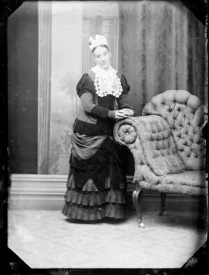 Unidentified woman, leaning on a chaise longue and wearing ornate headgear and a lace bib, a grey bodice and pleated skirt, both with dark trim