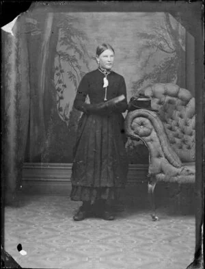 Unidentified woman, standing by a chaise longue, with a book held in both hands, and wearing a plain dark bodice and patterned skirt, with [lace-up?] shoes