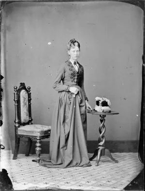 Unidentified woman, wearing a skirt and embroidered bodice and holding a [riding?] whip, hat and gloves on the nearby table