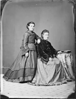 Unidentified woman with hair in a snood and wearing a full length skirt and bodice with elaborate long cuffs, is leaning on the back of the chair in which another unidentified woman is sitting, wearing a full skirt in a checked material and a black velvet jacket with elaborate cuffs