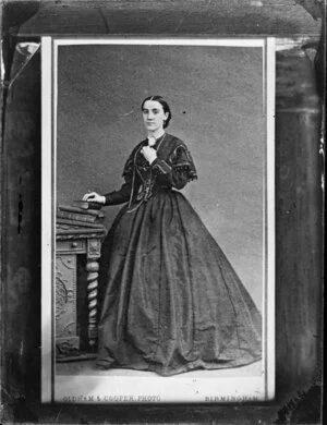Unidentified woman, wearing a patterned dress with a full skirt and silver trim on the bodice and standing by a small table piled with large volumes