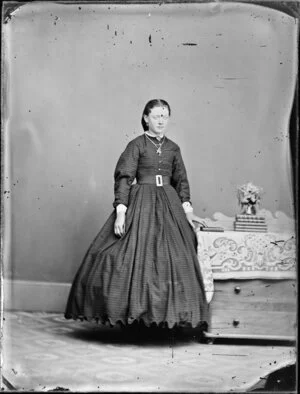 Unidentified woman, wearing a full-skirted dress with horizontal stripes and standing at a table with books