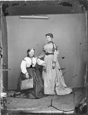 Unidentified young Maori woman, standing with a hand on the shoulder of a seated woman, holding a kete/flax basket