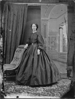 Unidentified woman, wearing a bodice with full sleeves over a full skirtand resting a hand onl the back of a chair