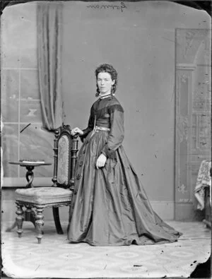 [Mrs?] Gorman, leaning on a chair back and wearing a fringed bodice and a very long, full skirt
