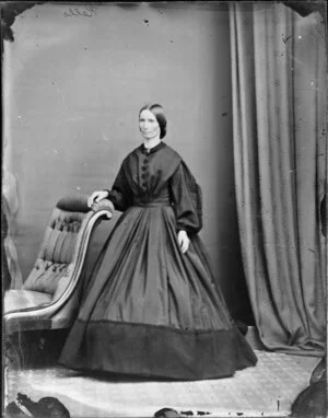 Mrs Kells, wearing a loose jacket and a long skirt in a contrasting colour and resting a hand on the back of a chair