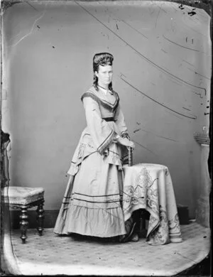 Unidentified woman, with bouffant hairstyle with ringlets, wearing an overdress and [bustle?l over full length skirt with a ruffle and holding a book