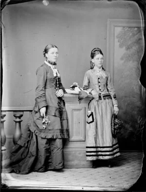 Two unidentified young women, both with intricate hairstyles, one wearing a full length dark skirt and a long bodice with ornate cuffs and the other wearing a striped skirt and a fringed bodice with insets
