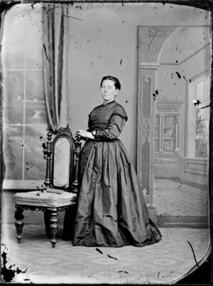 Unidentified woman, holdin the back of a chair and wearing a bodice with caped sleeves over a full skirt