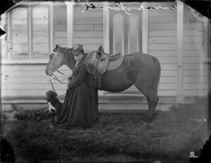 Mrs Anna Lower, wearing a dark bodice and skirt and a hat and standing by a saddled horse and a waiting dog