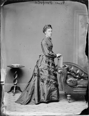 Miss Penfold, with hair piled on top of her head, wearing a close-fitting bodice and an intricately decorated skirt with a bustle and leaning on a book on the arm of a chaise longue