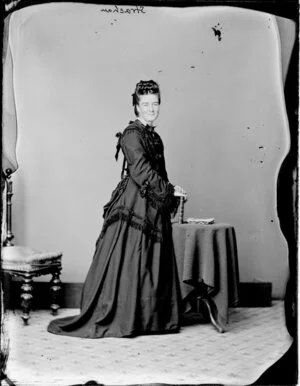Mrs Strachan, with hair piled on top of her head, wearing a fringed and beribboned dark bodice with cuffs and a full length skirt with a bustle