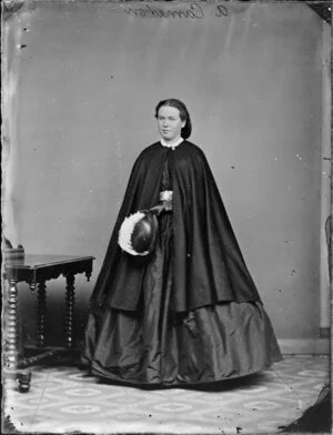 Mrs A Cameron, Holding a black hat with white feathers and wearing a dark cape over a full skirt with a metal buckle