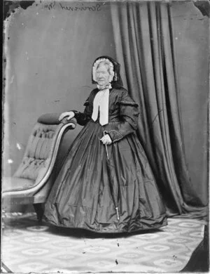 Mrs Scrivener, wearing a bonnet and a dress with decoration around cuffs and cape sleeves and holding an umbrella