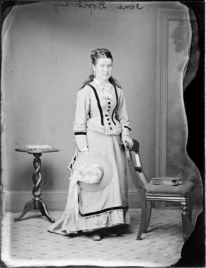 Jane Donovan with her hair coiled on top of her head, wearing a bodice with trims, over a long skirt with a frilled hem and holding a straw hat