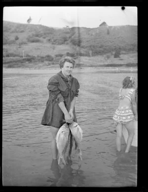 Unidentified woman standing in the sea and holding three large fish, Whitianga