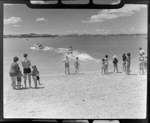 People enjoying a day at Maraetai beach, Auckland, including a water skier being towed out to sea in the background
