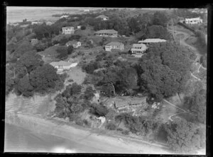 View of two houses being build on slopes of Karaka Bay hillside above beach, one with partially collapsed roof, amongst other residential houses with road and path to beach, Glendowie, Auckland City, with farmland beyond