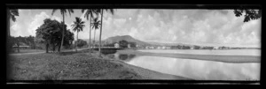 View in Samoa [probably near Apia], looking along a curved beach and river estuary towards houses at the bottom of a steep hill in the distance
