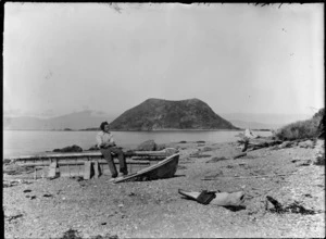 Beach scene with unidentified boy sitting on wreck of a dinghy, includes island in background, Marlborough Sounds
