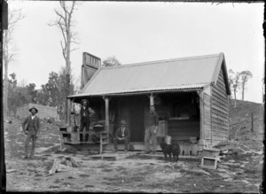 Unidentified group of men with dog, standing outside a wooden hut, which sits on recently cleared ground, possibly Wellington region
