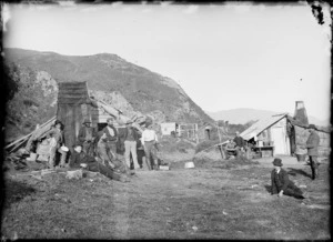 Unidentified group of men from a construction gang, sitting outside a group of huts, at the base of a hill, [Marlborough Sounds?]