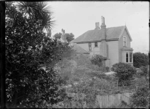 View across suburban gardens towards a two-storied wooden house with brick chimneys, possibly Wellington region