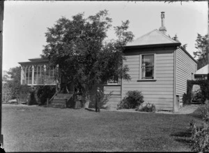 An unidentified man and woman standing beneath a tree outside a single-storied wooden house, which has a sunrooom and a corrugated iron roof, location unidentified
