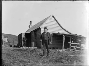 Unidentified man in a bowler hat, standing with a boy outside a wooden cottage with a shingled roof and a lean-to, location unidentified