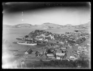 View from hill above Port Chalmers wharves, overlooking wharf buildings, part of the township near the wharves, part of Otago Harbour, and an unidentified steam ship moored at the wharf.
