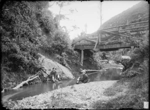 Photographer William Williams and an unidentified man, who is smoking a pipe, sitting on the banks of Kaiwharawhara Stream, Wellington Region, including derelict bridge