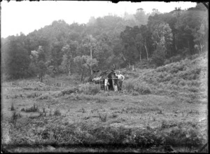 Thomas H Wyatt with pack leaning on a small hut at the edge of the bush, unidentified location
