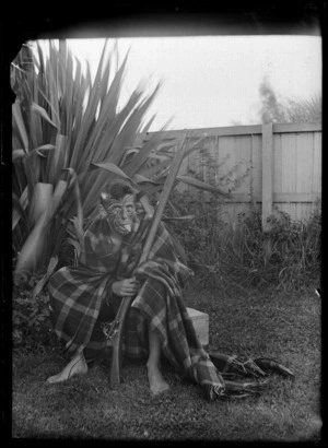 Seated unidentified man with his tongue stuck out, wearing a blanket, holding a rifle and with his face painted to look like a Maori tattoo, in the back yard of the 'Old Shebang', Cuba Street, Wellington