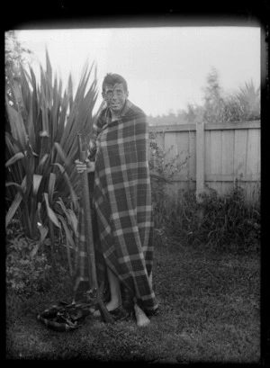 Unidentified man wearing a blanket, holding a rifle and with his face painted to look like a Maori tattoo, in the back yard of the 'Old Shebang', Cuba Street, Wellington