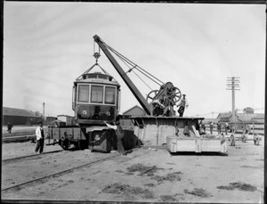 Men using a crane to load a tram for Whanganui, onto a train carriage, Christchurch railyards