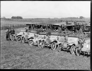 World War I invalided troops arrive home, transported in cars lined up in three rows on a field, front row cars open topped with two men standing along side looking on, farmland beyond