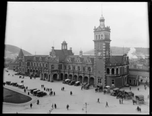 Dunedin Central Railway Station with people, cars and horse drawn wagons on forecourt, with commercial and residential buildings behind, Otago Harbour and Peninsula beyond