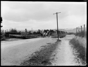 Horse drawn coach with men in suits on dirt road, crossing Pahiatua Bridge on the outskirts of Pahiatua, Tararua District, wooden bridge with advertising 'use Clapham's patent horse holder' in white paint on timbers, cleared land with tree trunks beyond