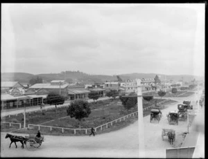 Pahiatua main street, Tararua District, view from upper floor building of Main Street showing fenced grass median strip street divider with people on horse drawn carts, with local businesses Milnes Central Book Arcade, C Sanford & Co - Drapers & Clothing, cleared hill land behind town