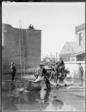 Firemen on training exercise showing water pumping apparatus in action, with hoses and fire truck, with senior officer looking on, Christchurch Fire Brigade