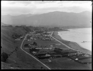 Seaside resort town of Kaikoura showing the Esplanade and Torquay Street with houses and fields, commercial buildings and primary school mid-view, farmland and seaward Kaikoura Mountain Range beyond